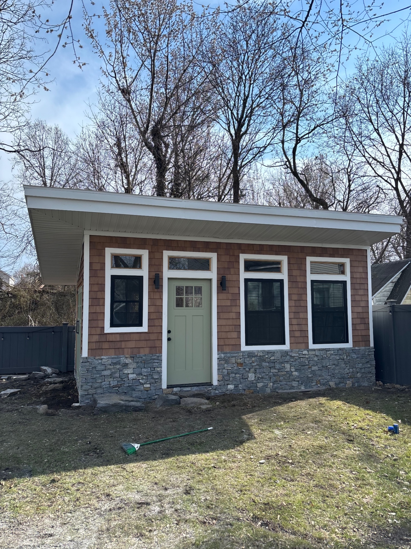 Finished backyard cabin with cedar shake siding, stacked-stone veneer base, sage green craftsman door, large black-trim windows, and modern shed roof