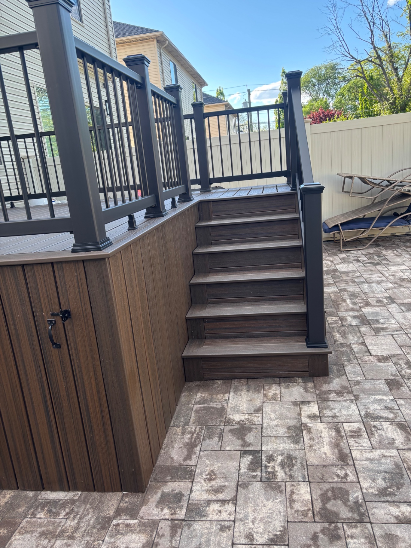 Close-up of finished composite deck stairs with brown treads, vertical skirting with hidden storage door, and black aluminum railings