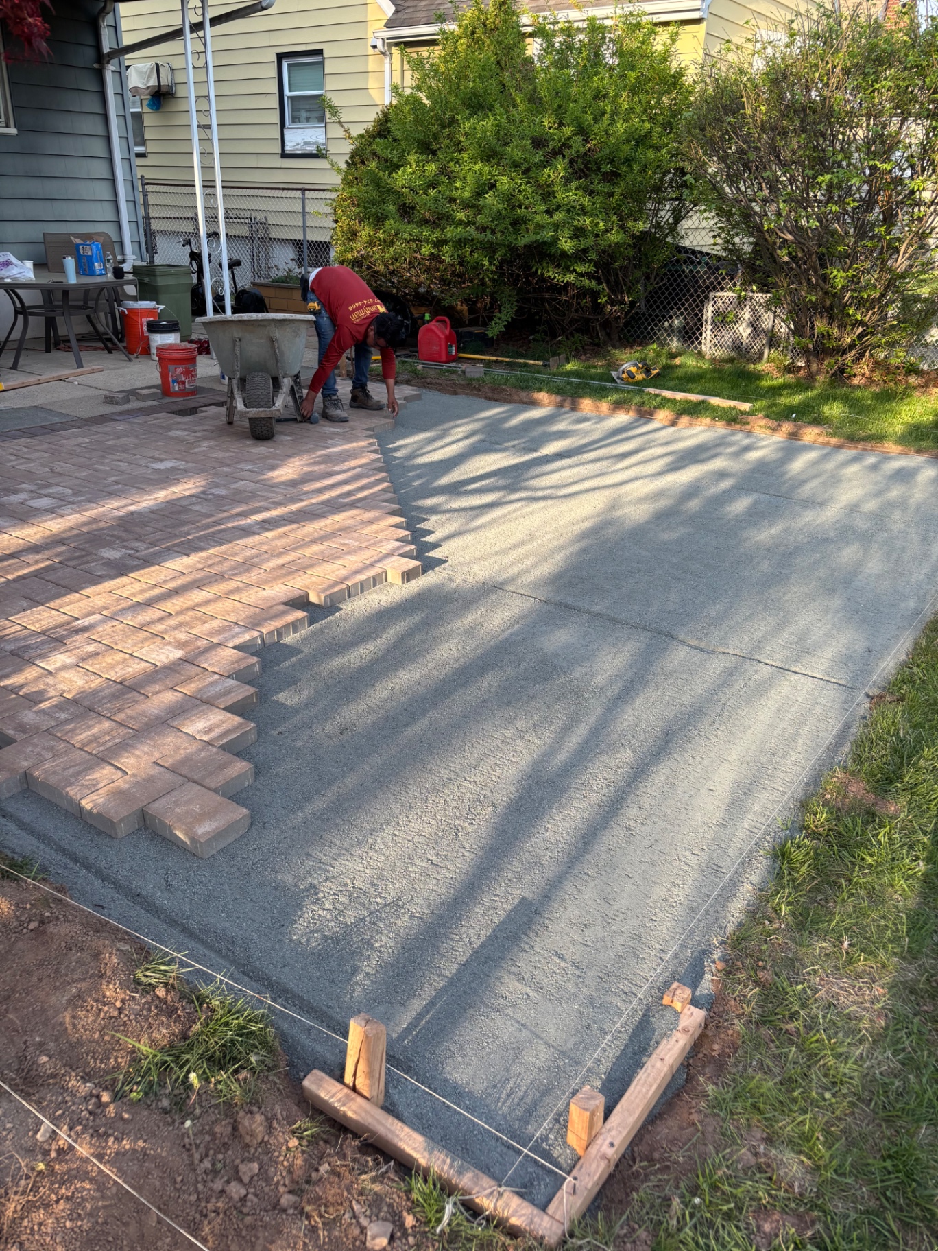 Paver patio installation in progress with crew laying interlocking pavers over compacted base alongside fresh concrete pad
