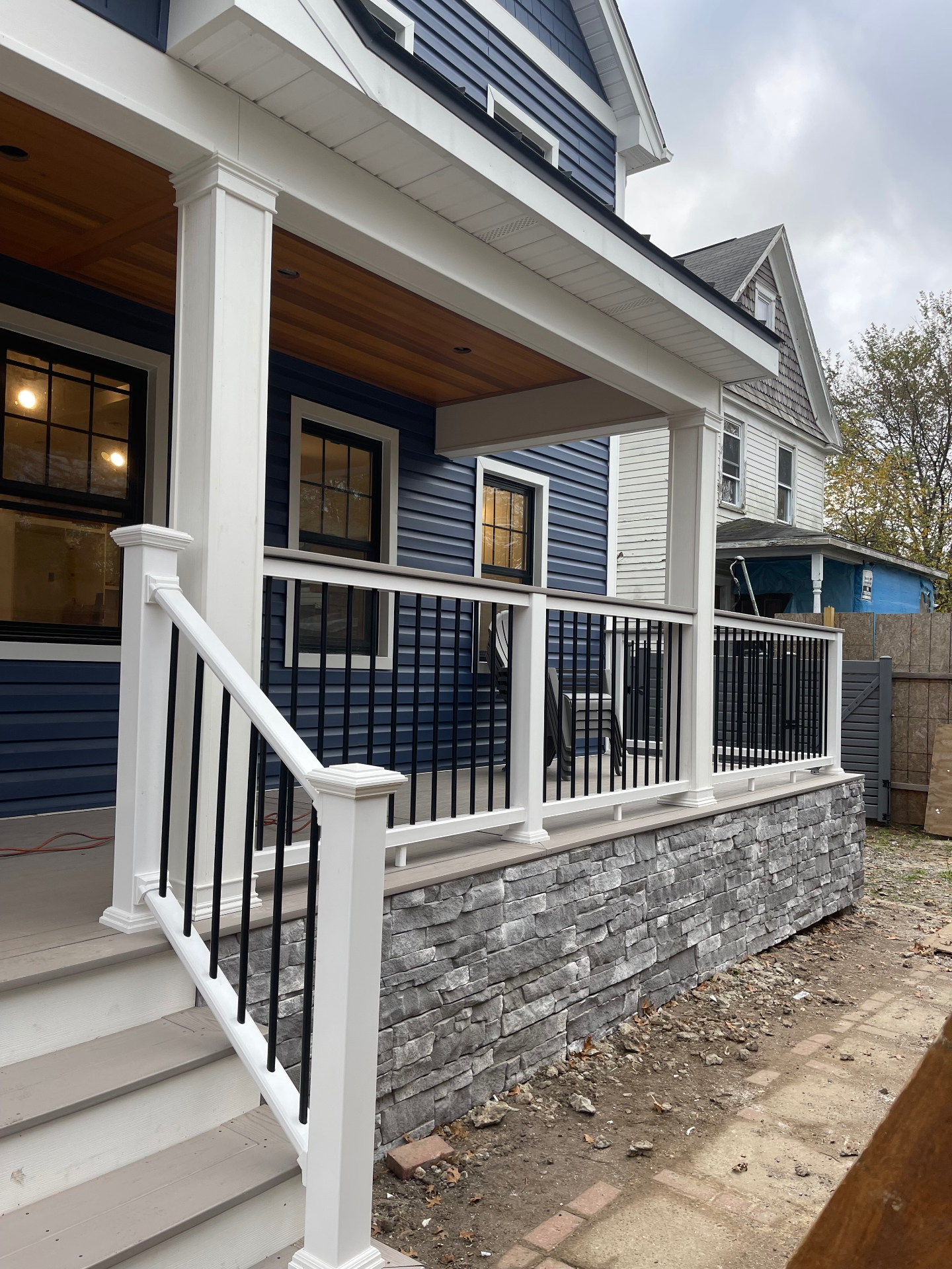 Finished covered front porch with white columns and railings, black balusters, wood-look ceiling, composite steps, and stacked-stone veneer base